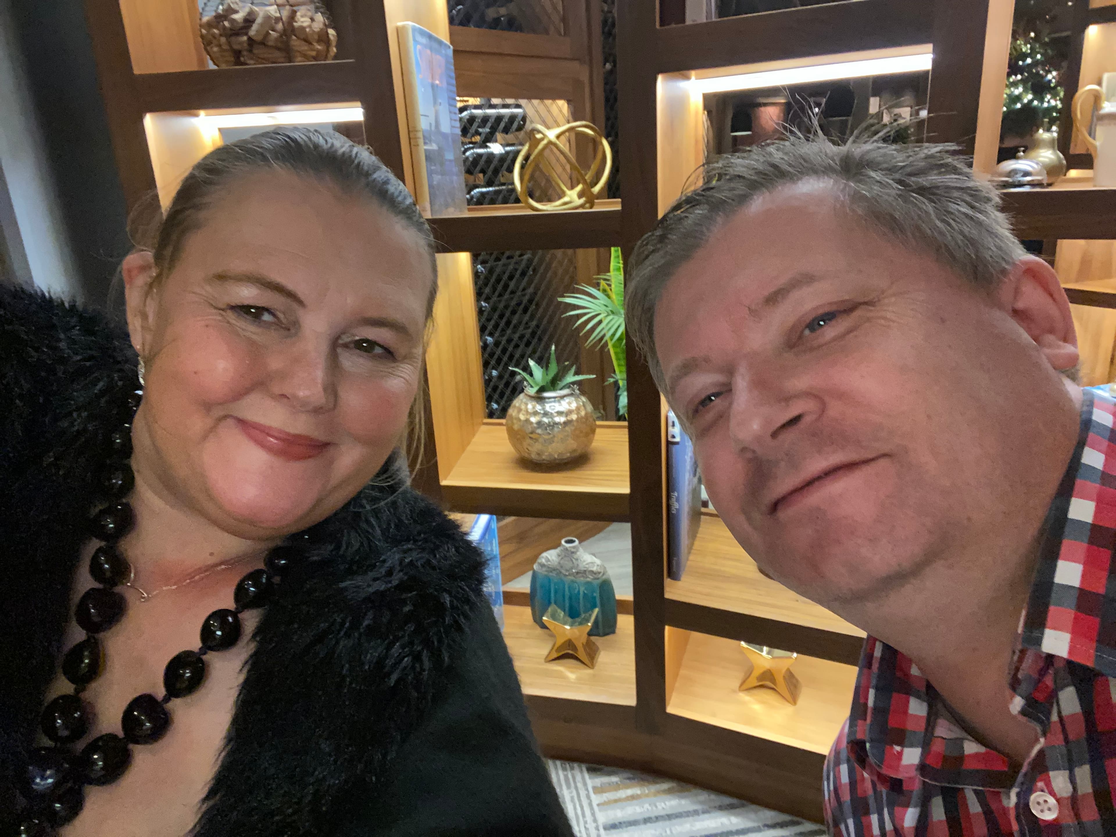 Smiling man and woman take a selfie in front of a warm lit wooden bookshelf.
