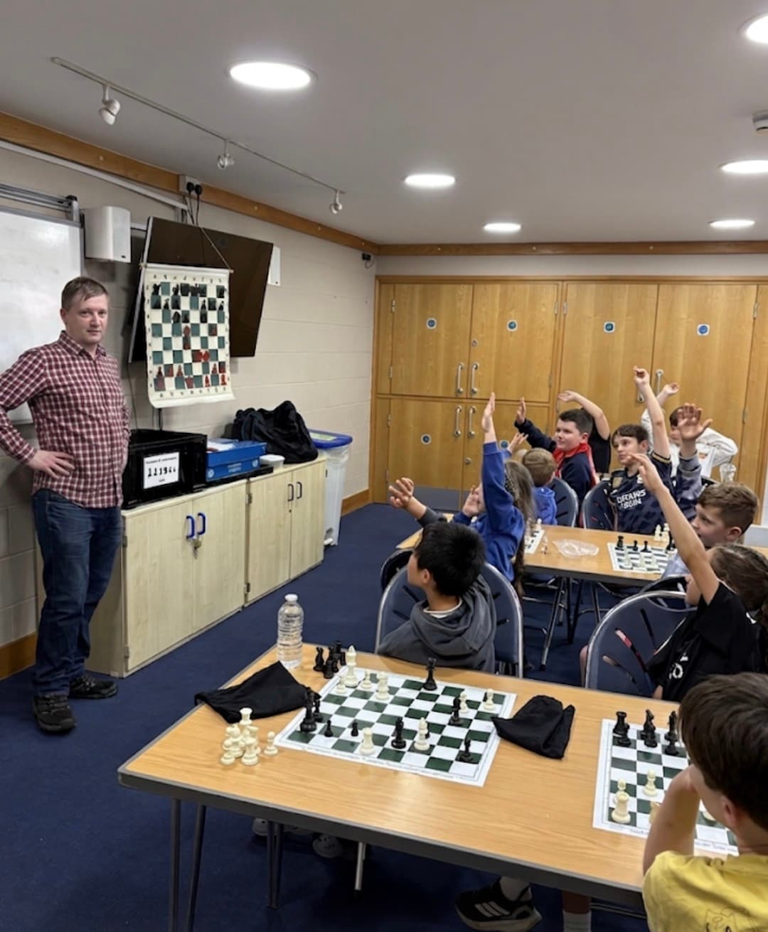 A chess instructor stands by a demonstration board as children raise their hands.