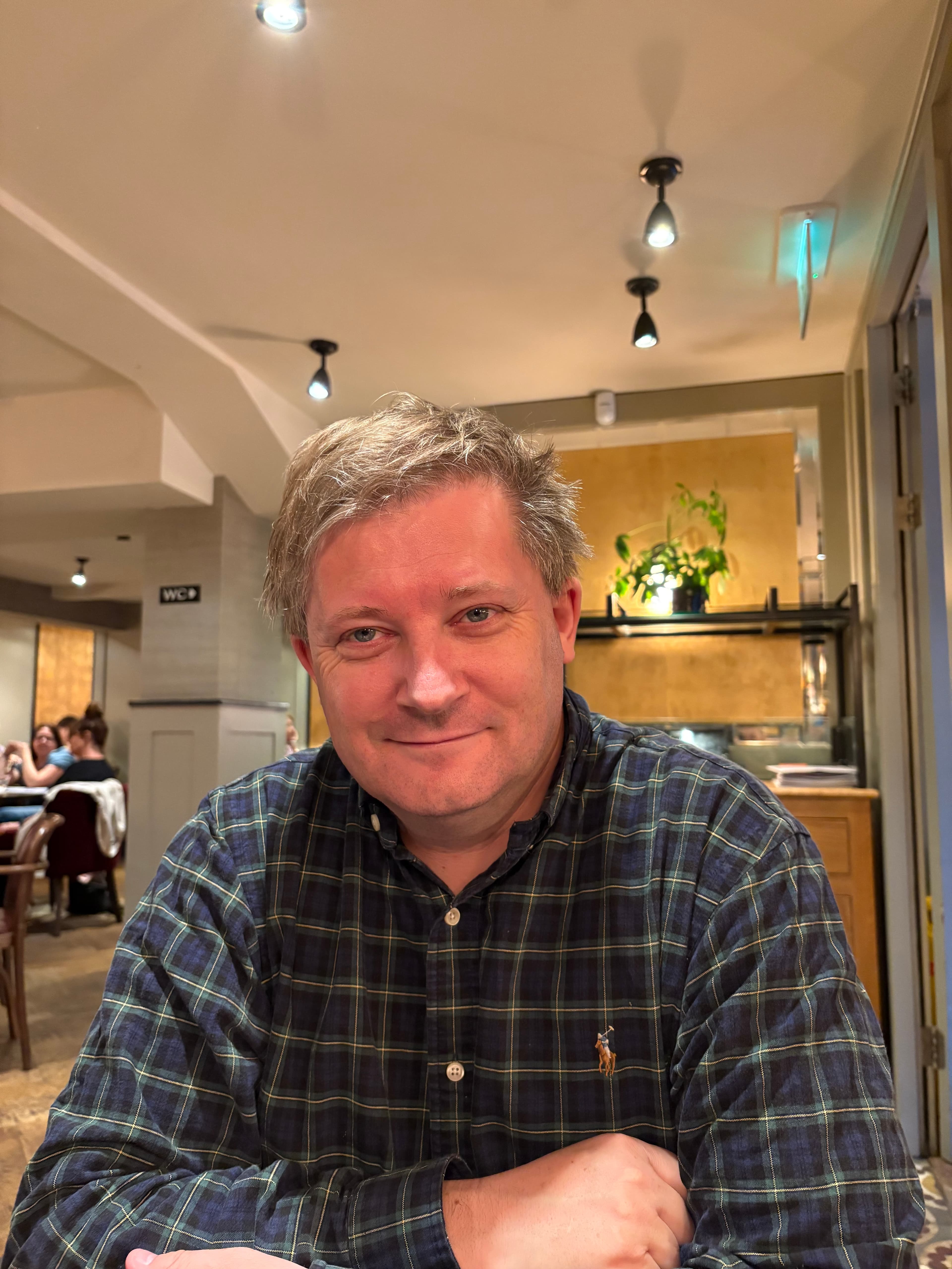 Smiling man with messy blonde hair wearing a plaid shirt in a warmly lit restaurant.